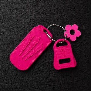 May include: A bright pink keyring set featuring a rectangular tag with three embossed claw marks, a smaller bottle opener attachment, and a flower-shaped charm, all connected by a silver-toned metal ball chain on a textured black surface.