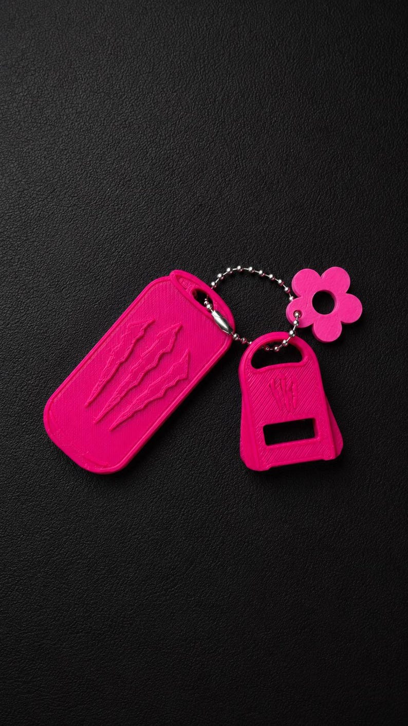 May include: A bright pink keyring set featuring a rectangular tag with three embossed claw marks, a smaller bottle opener attachment, and a flower-shaped charm, all connected by a silver-toned metal ball chain on a textured black surface.