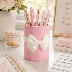 Puede incluir: Un portalápices cilíndrico de color rosa con un llamativo lazo decorativo blanco envuelto alrededor del centro. El recipiente contiene varios bolígrafos en tonos rosa, blanco y oro metálico, colocados verticalmente sobre una superficie.