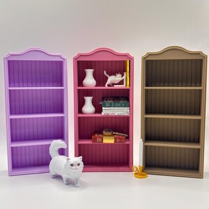 Puede incluir: Tres estanterías en miniatura en morado, rosa y beige. La estantería rosa está decorada con jarrones blancos, una figura de gato blanco, libros y una llave. Una figura de gato blanco está delante de la estantería morada. Un candelabro amarillo está delante de la estantería beige.