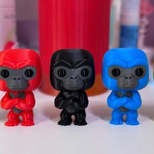 K&ouml;nnte beinhalten: F&uuml;nf kleine Gorillafiguren stehen in einer Reihe, jede in einer anderen Volltonfarbe: leuchtendes Gr&uuml;n, kr&auml;ftiges Rot, mattes Schwarz, tiefes Blau und sonniges Gelb. Jede Figur hat ein dunkelgraues Gesicht, gro&szlig;e Augen und verschr&auml;nkte Arme.