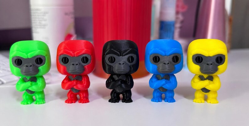K&ouml;nnte beinhalten: F&uuml;nf kleine Gorillafiguren stehen in einer Reihe, jede in einer anderen Volltonfarbe: leuchtendes Gr&uuml;n, kr&auml;ftiges Rot, mattes Schwarz, tiefes Blau und sonniges Gelb. Jede Figur hat ein dunkelgraues Gesicht, gro&szlig;e Augen und verschr&auml;nkte Arme.