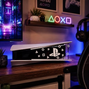 May include: A white and black gaming console faceplate featuring the PlayStation logo and iconic triangle, circle, cross, and square controller symbols. The custom cover is designed to fit over the central black section of the console hardware.