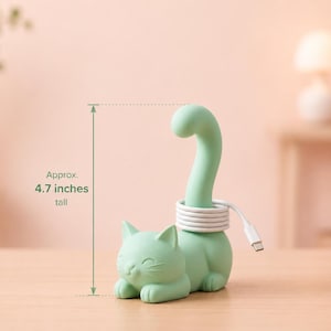 May include: A light green cat-shaped cable organizer with a tall, curved tail used for wrapping a white charging cord. The figure features a resting cat with closed eyes and pointed ears. The total height is approximately 4.7 inches tall.