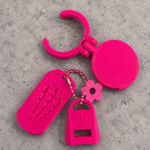 May include: A bright pink keychain set featuring a circular clip, a rectangular tag with three claw mark indentations, and a smaller trapezoidal tag with a cutout and a small flower charm, all connected by a matching pink ball chain.