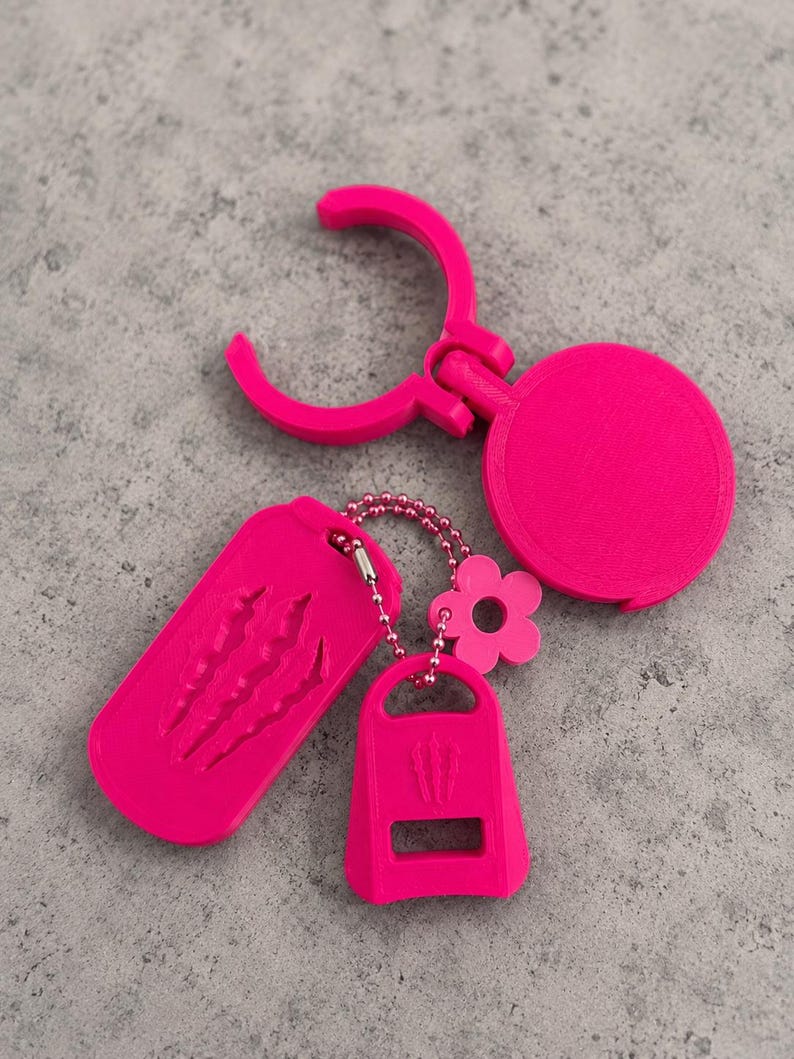 May include: A bright pink keychain set featuring a circular clip, a rectangular tag with three claw mark indentations, and a smaller trapezoidal tag with a cutout and a small flower charm, all connected by a matching pink ball chain.