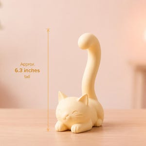 May include: A pale yellow cat-shaped figurine with a long, upright tail. The cat is in a resting position with its front paws tucked forward. A measurement graphic indicates the object is approximately 6.3 inches tall.