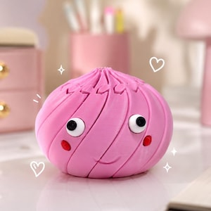 May include: A pink, cartoon-style dumpling with a smiling face, black eyes, and red cheeks. The dumpling has a textured surface and is set against a blurred background with other pink objects.