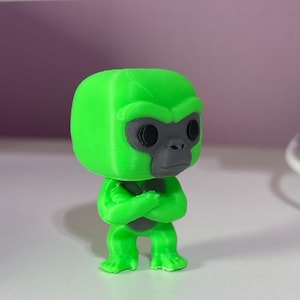 K&ouml;nnte beinhalten: Eine kleine, 3D-gedruckte Gorilla-Figur mit einem leuchtend neongr&uuml;nen K&ouml;rper und einem kontrastierenden dunkelgrauen Gesicht. Die Figur hat einen gro&szlig;en, abgerundeten Kopf, schwarze kreisf&ouml;rmige Augen und verschr&auml;nkte Arme vor der Brust.