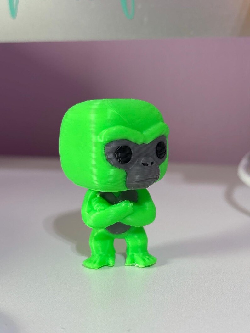 K&ouml;nnte beinhalten: Eine kleine, 3D-gedruckte Gorilla-Figur mit einem leuchtend neongr&uuml;nen K&ouml;rper und einem kontrastierenden dunkelgrauen Gesicht. Die Figur hat einen gro&szlig;en, abgerundeten Kopf, schwarze kreisf&ouml;rmige Augen und verschr&auml;nkte Arme vor der Brust.