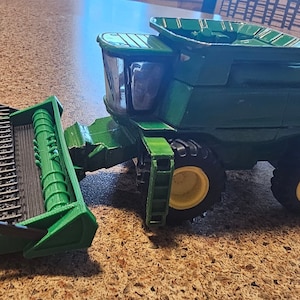 May include: A green and yellow toy combine harvester with large black tyres. The combine has a detailed front cutting platform and a cab with a clear window. The toy is likely a replica of a John Deere model.