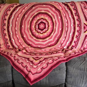 Könnte beinhalten: Eine große, kreisförmige Häkeldecke mit einem komplexen Mandalamuster in verschiedenen Rosatönen, die von hellem Pastell bis zu tiefem Magenta und Burgunderrot reichen. Das Design umfasst konzentrische Ringe aus strukturierten Maschen.