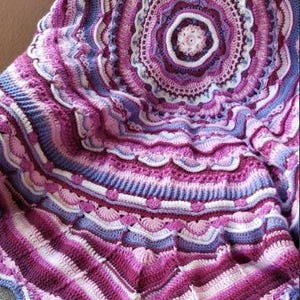 Könnte beinhalten: Eine große, gehäkelte Decke in den Farben Lila, Rosa und Weiß. Die Decke hat ein kreisförmiges Design mit einem gewellten Rand.