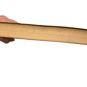 May include: A thick, old book with yellowed pages and a dark brown cover is held in a hand. The book's pages are visible from the side, showing their age and use. The background is plain white.