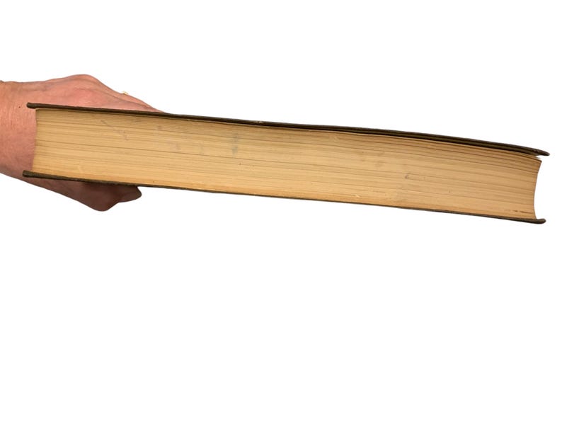 May include: A thick, old book with yellowed pages and a dark brown cover is held in a hand. The book's pages are visible from the side, showing their age and use. The background is plain white.