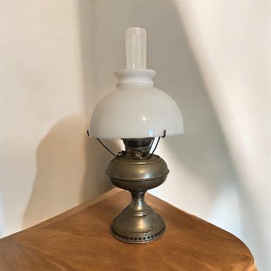 May include: An antique oil lamp with a white glass shade and a clear glass chimney. The lamp has a metal base and a decorative design around the bottom. The lamp is sitting on a wooden surface.