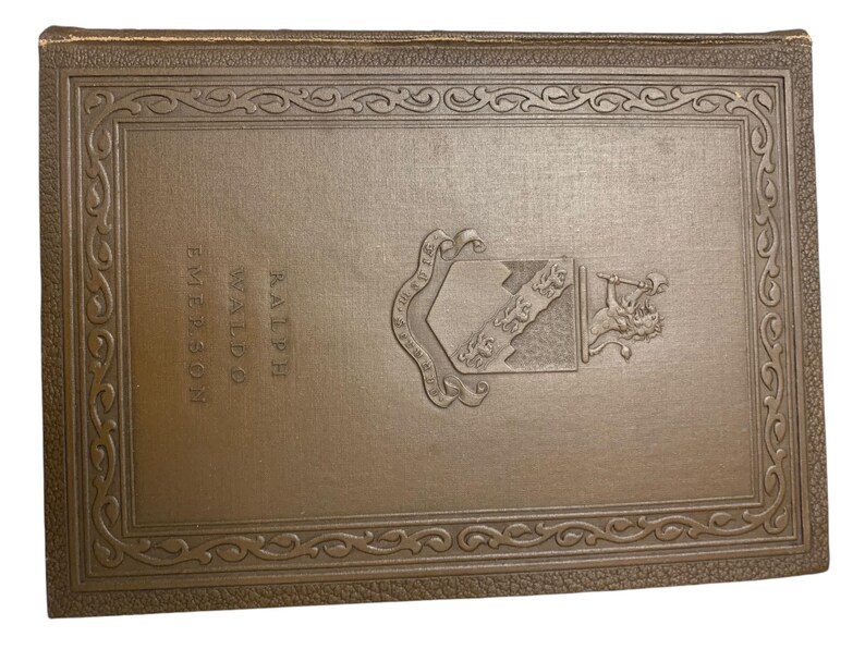May include: A vintage book with a brown cover featuring embossed text and a decorative border. The book's title is "Ralph Waldo Emerson". A detailed emblem is embossed on the cover, adding to its antique aesthetic.