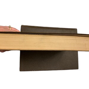 May include: A thick, vintage book with aged, yellowed pages and a dark brown cover. The book is held open, revealing the side profile of the pages. Another book is beneath it, also with a dark brown cover.