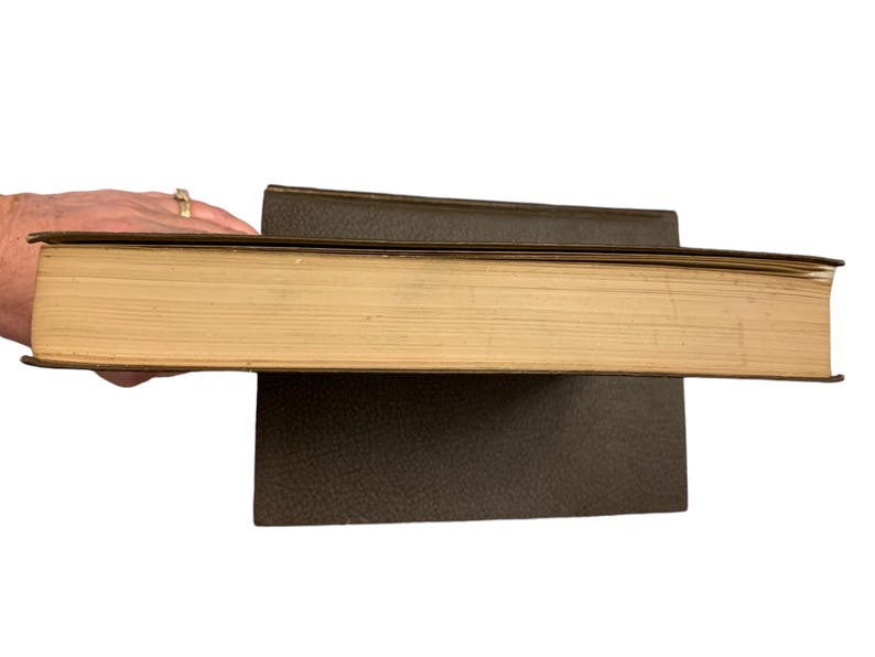 May include: A thick, vintage book with aged, yellowed pages and a dark brown cover. The book is held open, revealing the side profile of the pages. Another book is beneath it, also with a dark brown cover.