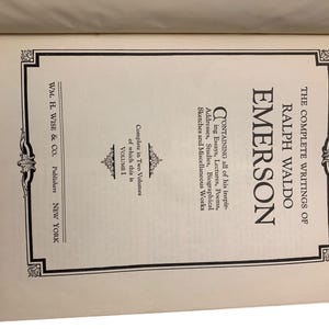 May include: Open book displaying the title "The Complete Writings of Ralph Waldo Emerson, Volume 1." The cover is brown, and the pages are cream-colored with black text. The book is published by WM. H. Wise & Co. of New York.