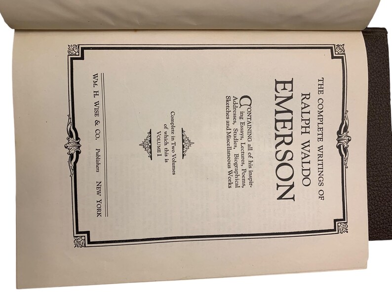 May include: Open book displaying the title "The Complete Writings of Ralph Waldo Emerson, Volume 1." The cover is brown, and the pages are cream-colored with black text. The book is published by WM. H. Wise & Co. of New York.