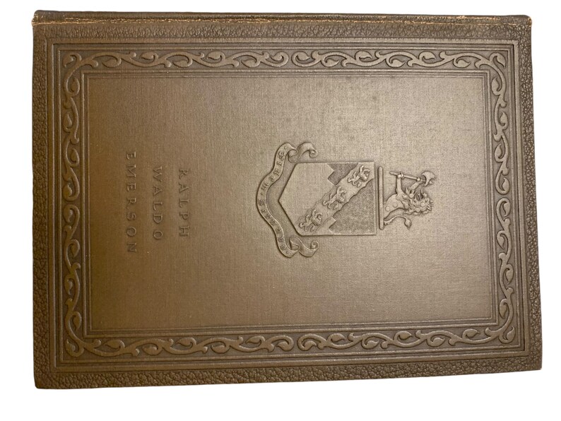 May include: A vintage book cover in a muted gold tone. The cover features embossed text reading "Ralph Waldo Emerson" and an ornate emblem. The border is decorated with a repeating scroll design, suggesting a classic or antique literary work.