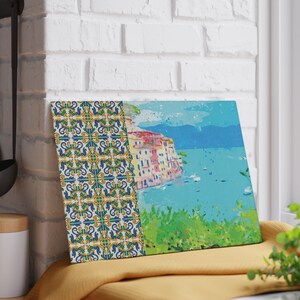 May include: A colorful painting of a seaside town with a blue tiled border. The town is built on a cliff overlooking the water. The water is a bright blue and there are boats in the distance.