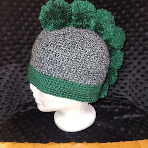 May include: Green and gray crocheted hat with a green pom-pom on top. The hat is on a white mannequin head.