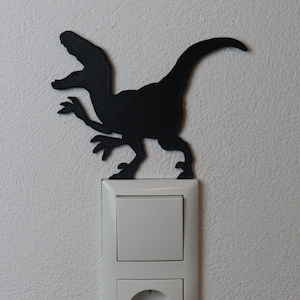 Könnte beinhalten: Eine weiße Steckdose und ein Lichtschalter mit einer schwarzen Dinosauriersilhouette oben drauf.