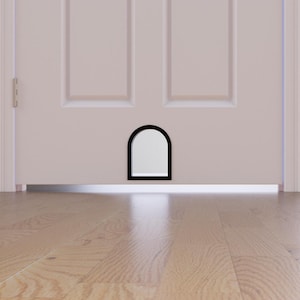 May include: A white door featuring a black-framed pet door. The pet door has an arched top and a rectangular shape. The door is set against a light-coloured wall and a wooden floor.