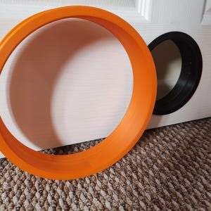 Könnte beinhalten: Ein leuchtend orangefarbener Ring mit leerem Zentrum, platziert in der Nähe einer weißen Oberfläche mit einer schwarzen kreisförmigen Öffnung. Das Objekt besteht aus einem festen Material, möglicherweise Kunststoff oder lackiertem Holz. Der Durchmesser beträgt ca. 15 cm.