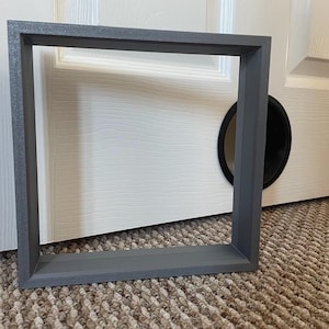 May include: A square, grey picture frame with a matte finish. The frame is empty, with no artwork or photo inside. The frame is placed on a patterned carpet in front of a white door with a black circular opening.