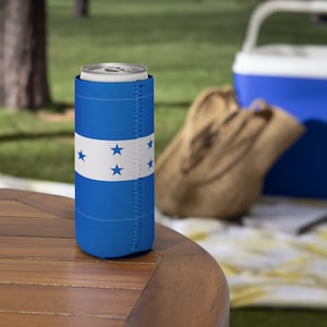 May include: A blue drinks can cooler displaying the Honduran flag. The design features a white band with five blue stars on a blue backdrop. The cooler sits on a wooden table, with a picnic scene visible in the background.