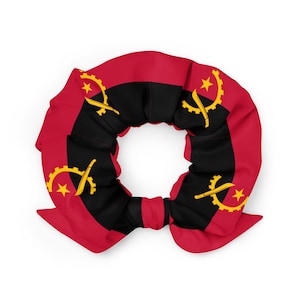 Op de afbeelding: Een scrunchie in de kleuren van de Angolese vlag: rood, zwart en geel. Het ontwerp bevat het embleem van de vlag, een tandwiel, een kapmes en een ster. De stof is geplooid en in een strikvorm geknoopt.