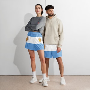 May include: Light blue and white shorts featuring the Argentinian flag design, with a sun emblem. The shorts are worn by two people, paired with a gray long-sleeve shirt and a beige hoodie. The shorts are made of a lightweight material.