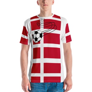 May include: White t-shirt featuring the Danish flag design with red rectangles and white crosses. A black and white football is depicted in motion, with curved lines trailing behind it. The shirt has short sleeves.