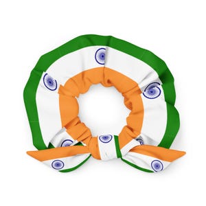 May include: A scrunchie featuring the colors of the Indian flag: orange, white, and green. The design includes the Ashoka Chakra, a navy blue wheel, repeated on the white sections. The scrunchie is tied in a bow.