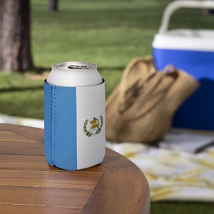 May include: A white aluminium can in a blue and white Guatemalan flag-themed can cooler. The cooler has a blue side with a stitched seam and a white side with the Guatemalan coat of arms. The can sits on a wooden table.