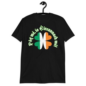 May include: Black t-shirt featuring a four-leaf clover design in the colors of the Irish flag: green, white, and orange. The text "Póg mé, is Éireannach mé!" arches above the clover. The shirt is hanging on a black hanger.
