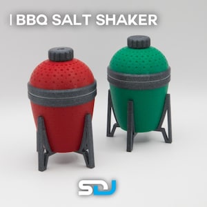 Op de afbeelding: Twee keramische zoutstrooiers in de vorm van barbecues. Eén is rood en de andere is groen. Beide hebben een zwarte basis met drie poten.