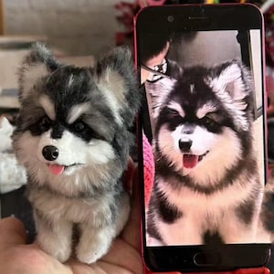 Puede incluir: Una figura de perro de peluche gris, negro y blanco, con una lengua rosa. La figura se sostiene en una mano junto a un teléfono inteligente que muestra una imagen similar. El pelaje del perro es suave y detallado.