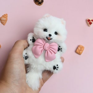 May include: A white fluffy dog toy with a pink bow, held in a hand against a pink background. The toy has black paw prints on its feet.