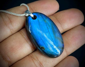 May include: A polished, oval-shaped labradorite pendant with a vibrant blue iridescence. The gemstone is set on a silver-colored bail and hangs from a delicate chain. The pendant is a striking piece of jewelry.