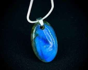 May include: Oval labradorite pendant with a silver chain. The polished gemstone displays vibrant blue and green hues. The pendant is suspended from a delicate silver chain, creating a striking contrast against the dark background. A beautiful piece of jewelry.
