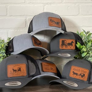 May include: Six gray and black trucker hats with brown leather patches featuring different designs of horse-drawn buggies. The patches include text such as "Amish Marketplace" and "Amish Uber".