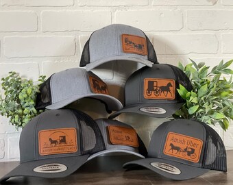 May include: Six gray and black trucker hats with brown leather patches featuring different designs of horse-drawn buggies. The patches include text such as "Amish Marketplace" and "Amish Uber".