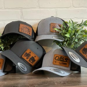 May include: A collection of trucker hats in various shades of gray and black. Each hat features a leather patch with unique designs and text, including trucks and the word "Truck". The hats have a mesh back and a snapback closure.