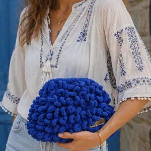 May include: A vibrant royal blue clutch bag featuring a dense, textured loop-knit surface. The bag has a rounded, pouch-like shape and includes a small, metallic gold-toned rectangular hardware accent on the lower right side of the exterior.