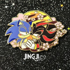 May include: Enamel pin featuring Sonic the Hedgehog and Shadow the Hedgehog embracing, set against a yellow and orange moon with cherry blossoms. The pin has a gold outline and the text "JINGJI ART".