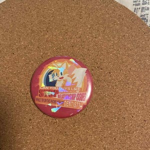 May include: A round, pink button with a holographic design featuring a cartoon fox character. The button has text that reads "CAN YOU GUYS LIKE SEO RELATIONSHIP ISSUES WHILE I'M ON THE PHONE WITH THE DENTIST?"